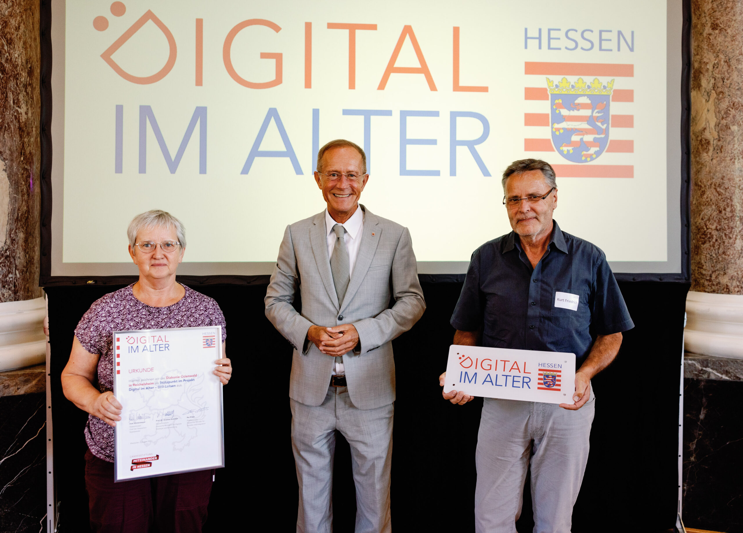 Staatsminister Axel Wintermeyer mit einer Mitarbeiterin der Diakonie und einem Di@-Lotsen stehen vor einer Leinwand